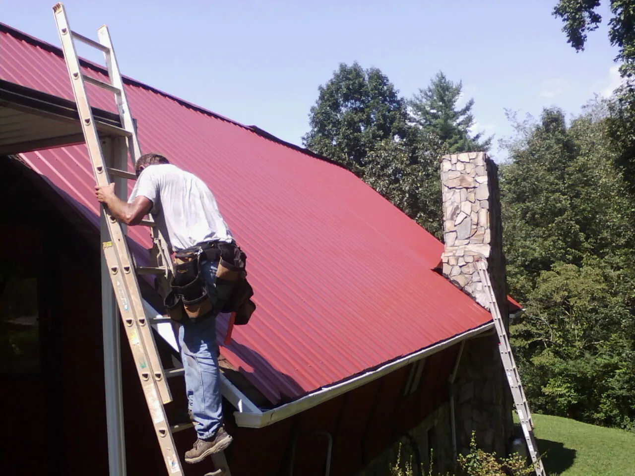 Roofer on a ladder beside a red metal roof and stone chimney — Soffit & Fascia Repair by Roofing Brothers in Willow Creek